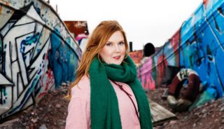Hannaleena Hauru kuvattuna graffitein maalatun veneen edessä. Päällä vihreä kaulahuivi ja vaaleanpunainen takki