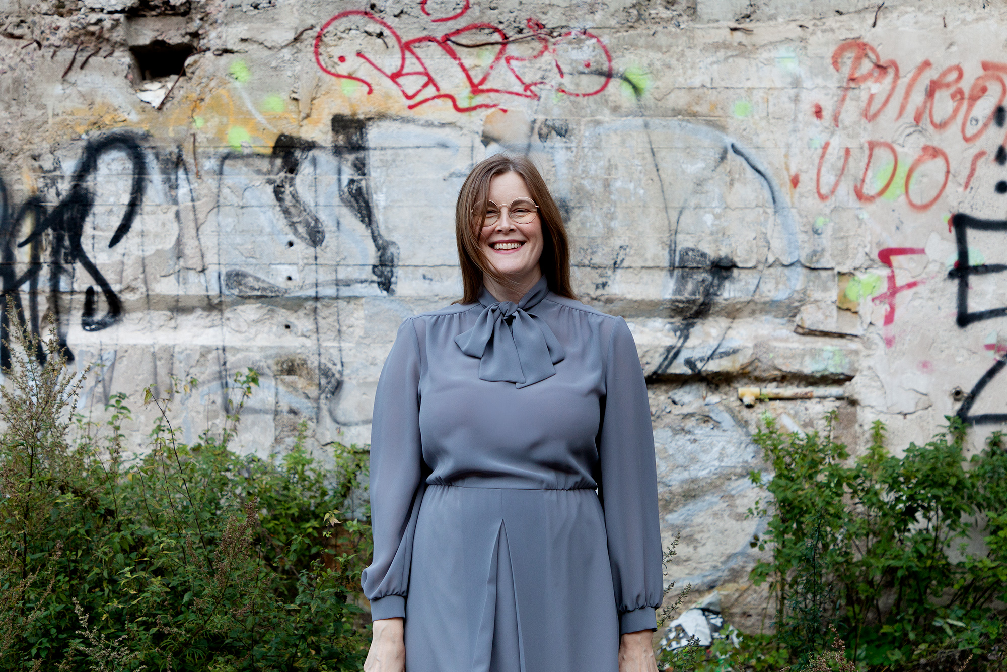 Aalto nauraa, katsoo kameraan, harmaa mekko päällään. Taustalla pensaita ja seinä, jossa graffiteja.