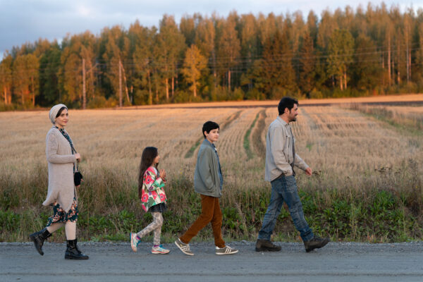 Pakolaisena Suomeen tulleet äiti, tytär, poika ja isä kävelemässä hiekkatietä. Taustalla pelto ja havumetsää.