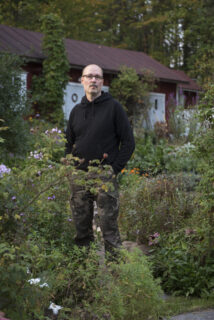 Kärkkäinen seisomassa puutarhassa kädet maastohousujen taskuissa. Musta huppari päällä. Taustalla punainen ulkorakennuksen seinä.