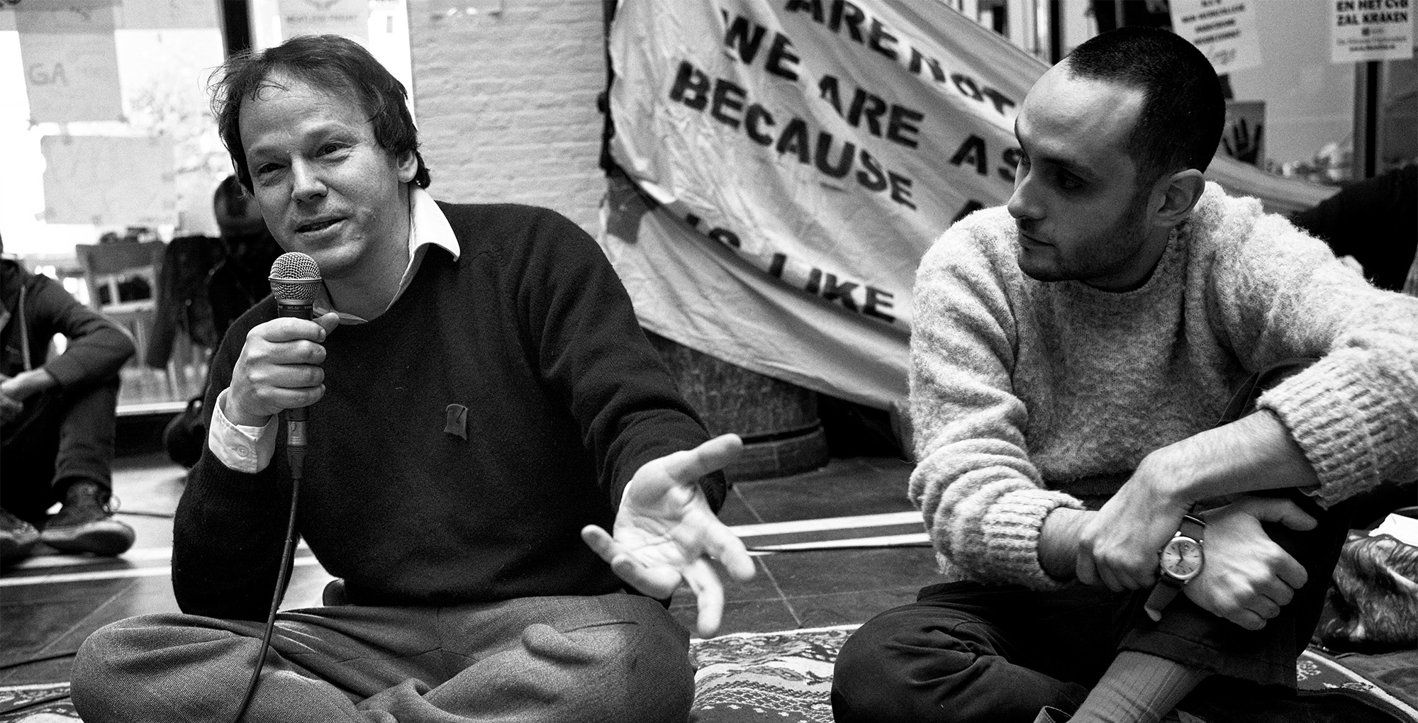 Graeber istuu toisen henkilön vieressä maassa, puhuu mikrofoniin.