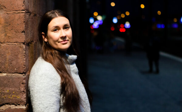 Tiina Elovaara katsoo kameraan hymyillen, Helsingin rautatieaseman laiturialueella valon alla. Taustalla pimeää.