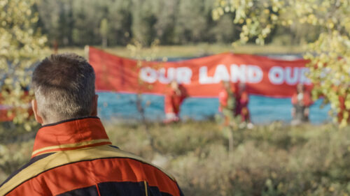 Saamenpukuisen henkilön niska kuvan etualalla, taustalla luontoa ja mielenosoittajia ja kyltti "our land our-".