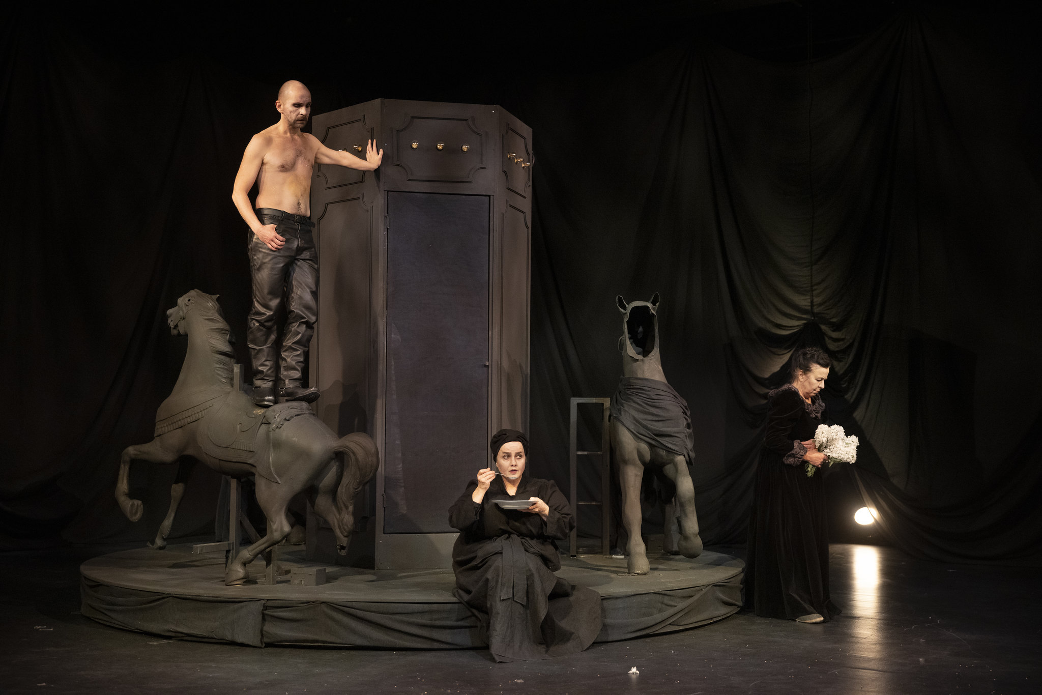 Kolme näyttelijää lavalla mustan lavasteen luona. Paidaton mies seisoo hevosen päällä ja nojaa lavasteeseen. Keskellä istuu nainen lautanen ja lusikka kädessä. Vasemassa reunassa vanhempi nainen kädessään valkoinen kukkapuska.