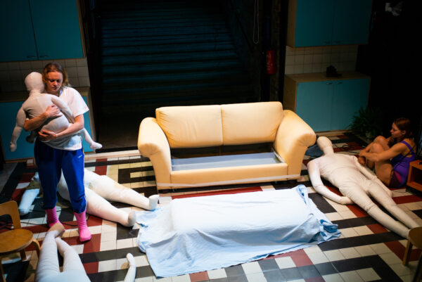 Näyttelijä halaa ihmisen kokoista nukkea. Keskellä sohva josta on otettu istuintyynyt irti. Oikeassa laidassa näyttelijä istuu maassa puristaen jalkoja rintaansa vasten.