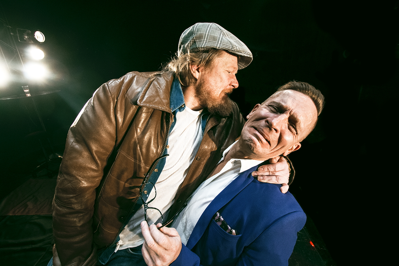 Kaksi henkilöä: nahkatakkiin pukeutunut pitelee pukuun pukeutuneen henkilön olkapäällä kättä. Pukuun pukeutunut itkee.