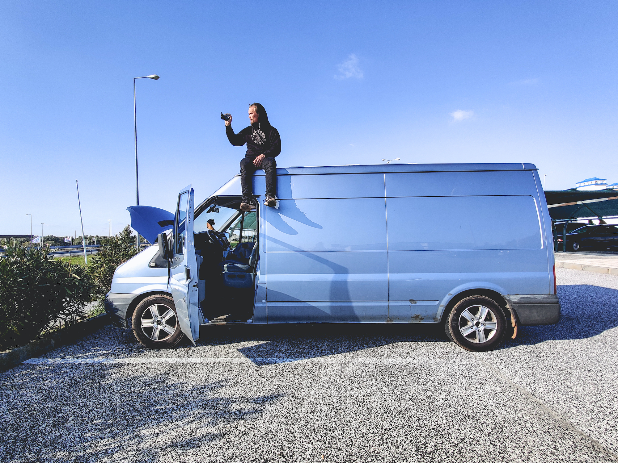 Mustaan huppariin pukeutunut mies kuvaa videokamarella vaalean sinisen pakettiauton katolla