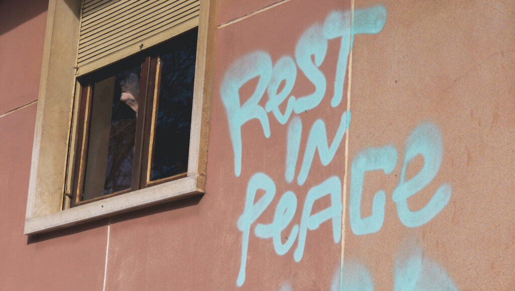 Vanha ihminen katsoo ulos ikkunasta. Ikkunan viereen vaaleanpunaiseen seinään on maalattu spraymaalilla teksti "Rest in Peace"