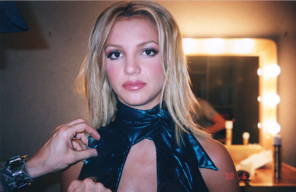 Britney Spears katsoo kameraan. Takana valaistu meikkipöytä. Kuvan ulkopuolelta tuleva käsi korjaa Britneyn mustaa asustetta.