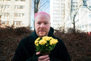 Haapa katsoo kameraan, pitelee käsissään keltaisia kukkia. Taustalla kerrostaloja.
