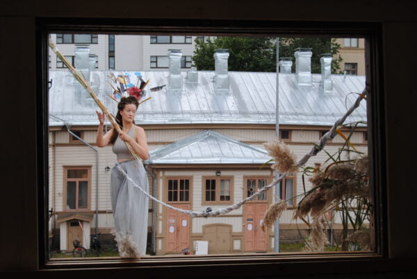 Kuvassa nainen ikkunalla, taustalla näkyy vanha puutalo.