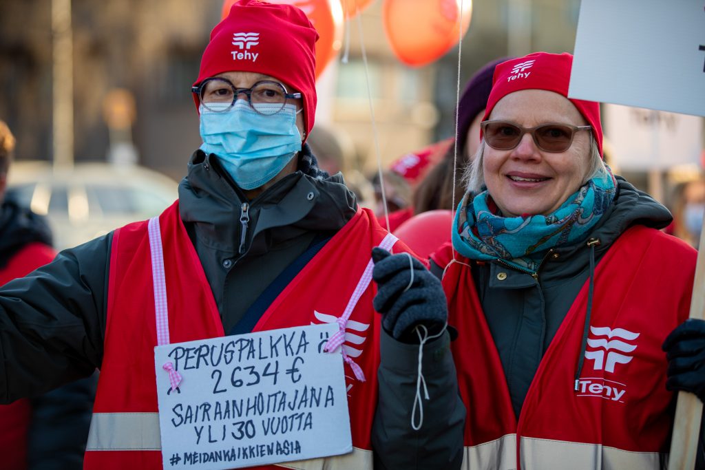 Kaksi mielenosoittajaa katsoo kameraan. Kyltissä lukee: "Peruspalkka 2634€ sairaanhoitajana yli 30 vuotta #meidänkaikkienasia".