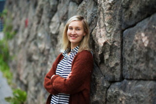 Anna Kivimäki-Pelluz nojaa kiviseinää vasten ja katsoo hymyillen kameraa