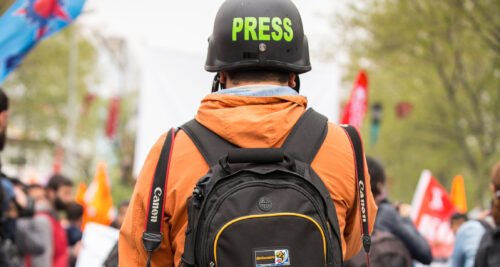 Kypäräpäinen henkilö valokuvattu takaapäin, olkapäillä roikkuu kameroita. Kypärässä lukee "press". Taustalla näkyy ihmisjoukko.