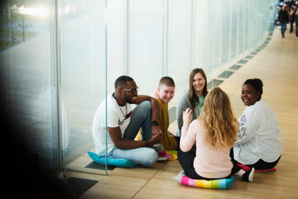 Viisi nuorta aikuista istuu tyynyjen päällä lattialla ja keskustelee toisilleen hymyillen