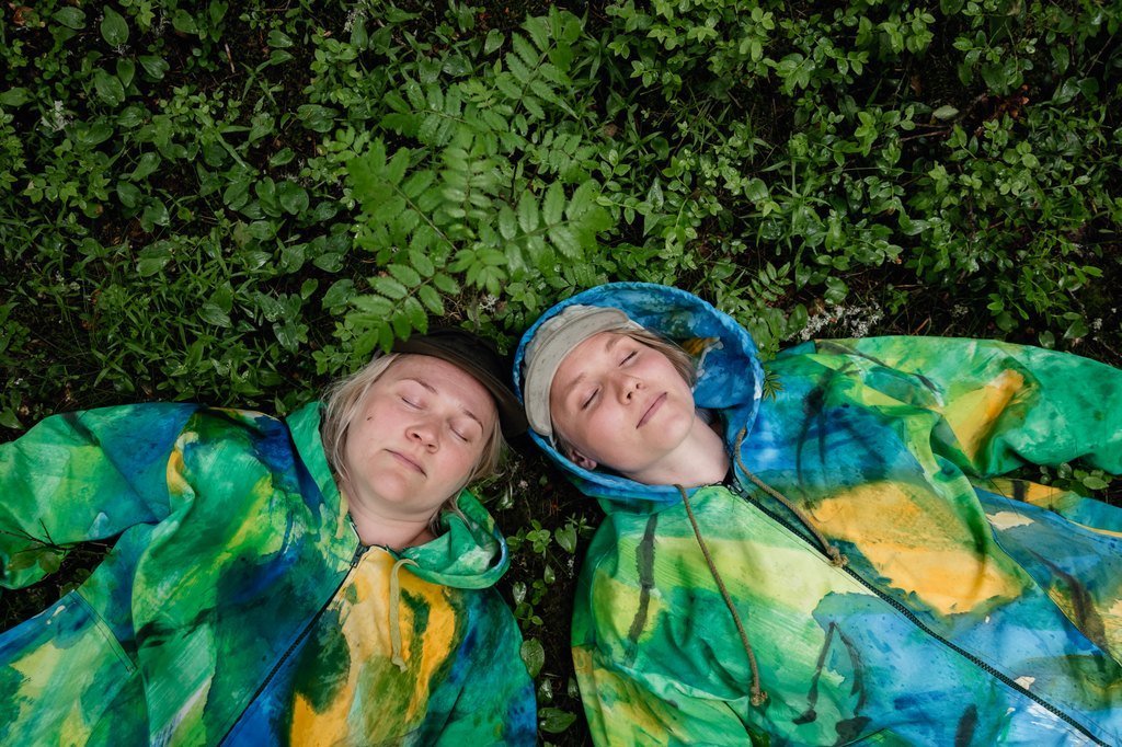 Kaksi henkilöä makaa maassa sadetakeissa, silmät kiinni.