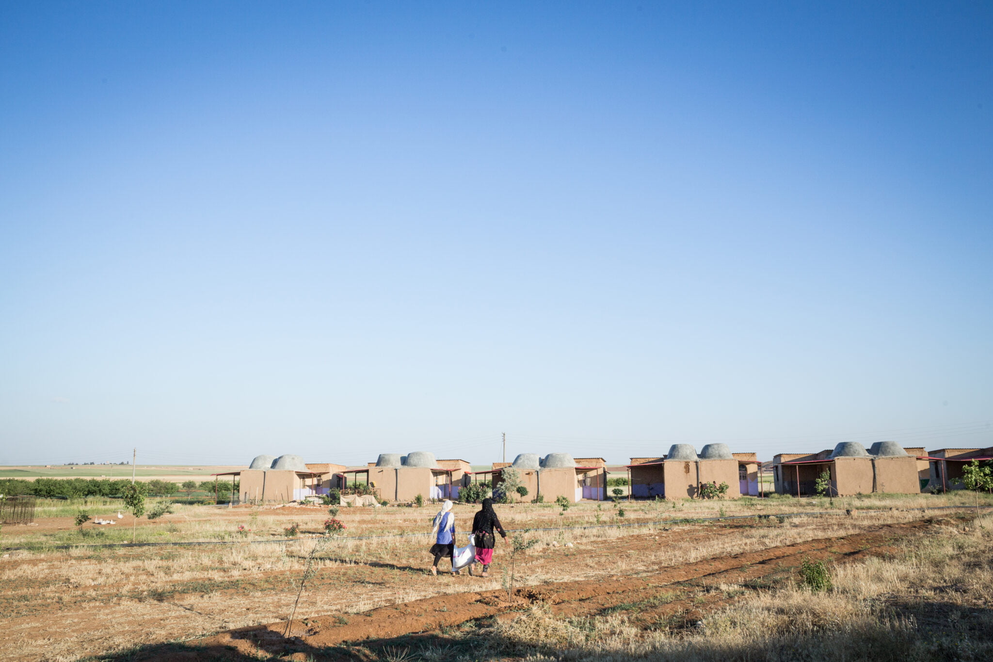 Kaksi naistaa kantaa säkkiä kohti kylää. Taustalla perinteisiä savesta rakennettuja rakennuksia.