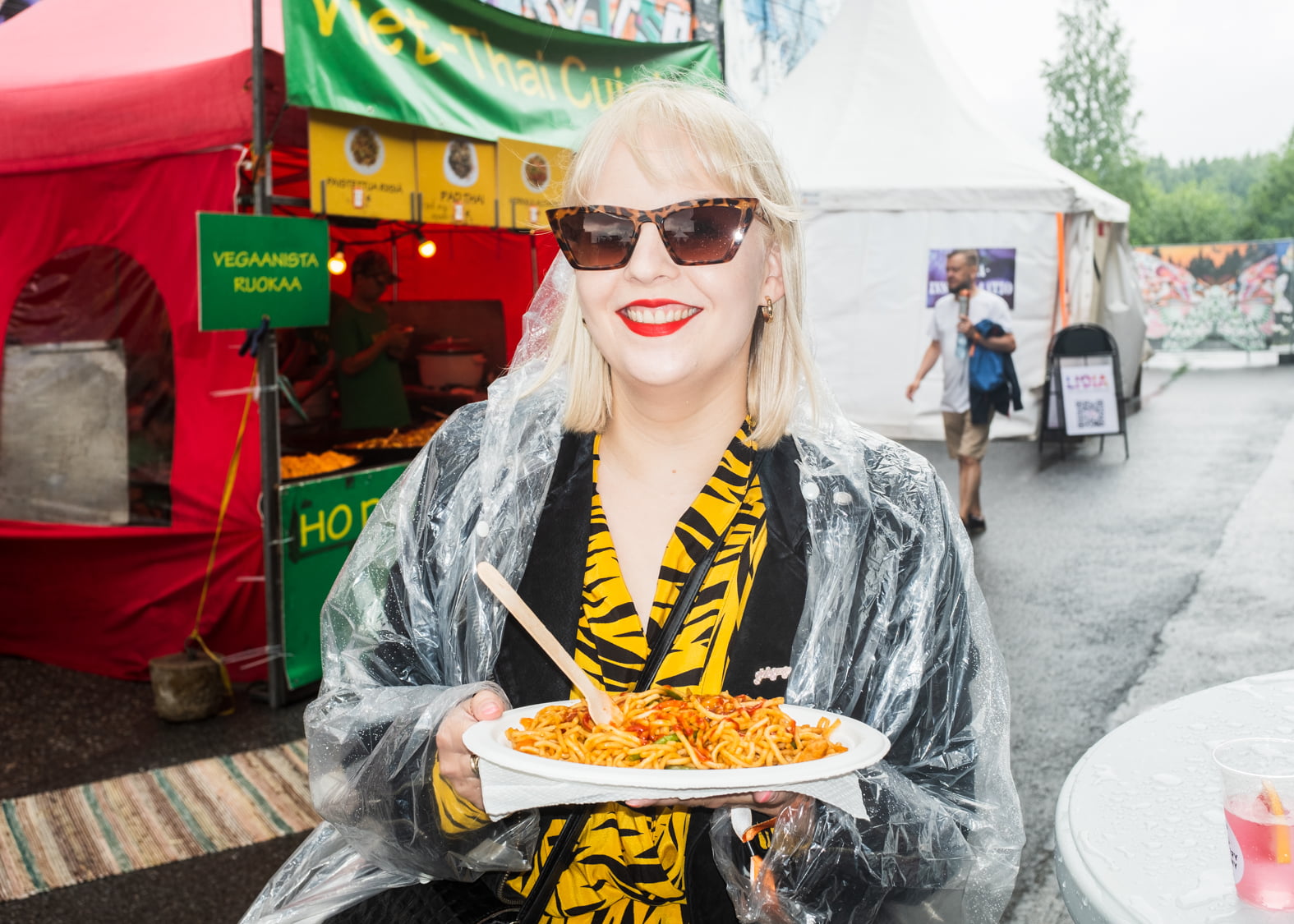 Vaaleahiuksinen nainen pukeutuneena kertakäyttöiseen sadetakkiin ja aurinkolaseihin hymyilee kameralle. Kädessä ruoka-annos.