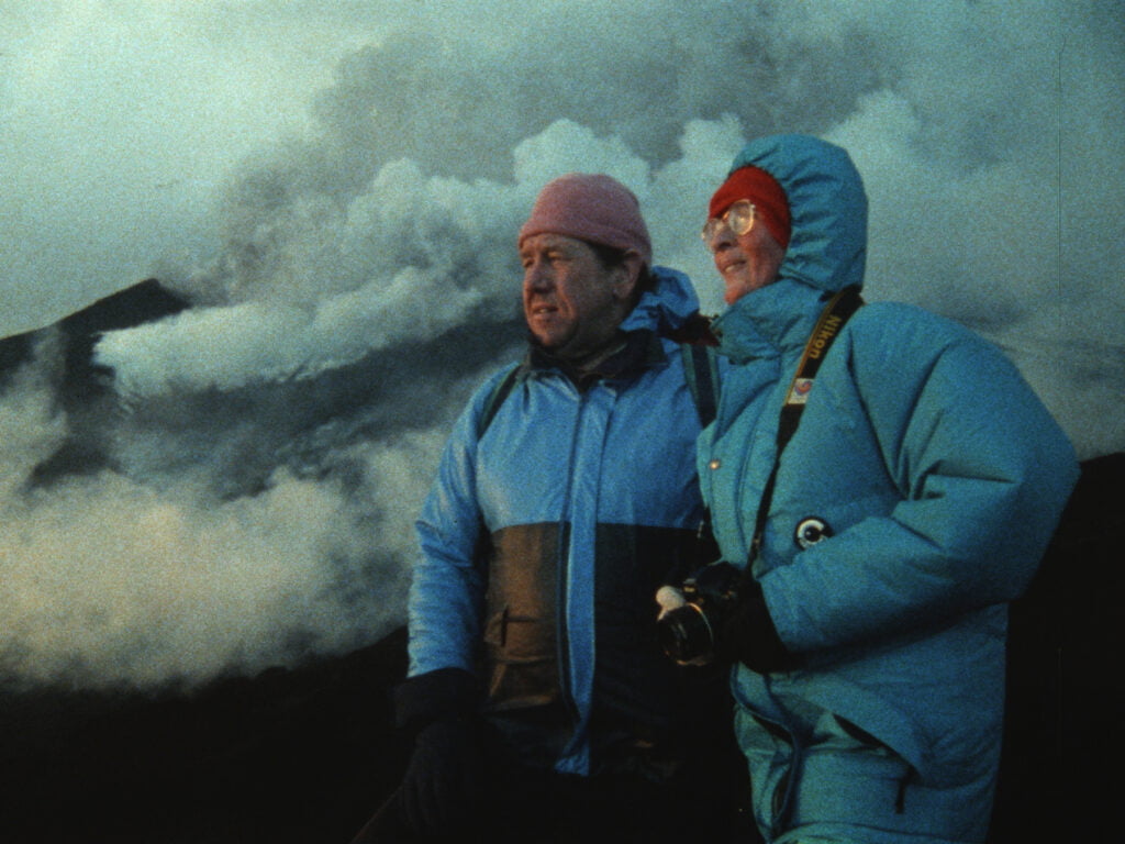 Kaksi henkilöä seisoo rinnatusten, taustalla vuoristoa ja savua.