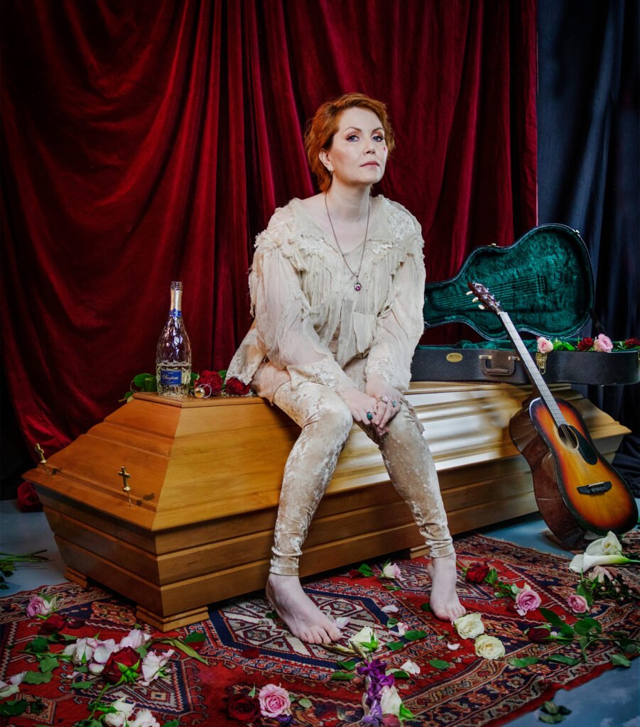 Astrid Swan istuu ruumisarkun päällä. Vieressä kitara, kuohuviiniä.
