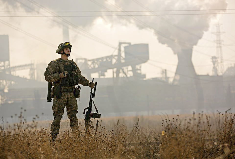 Sotilas seisoo pellolla, tastalla teollisuusrakennuksia.