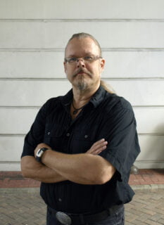 Markus Hotakainen katsoo kameraan, kädet puuskassa, tausatlla harmaa seinä.