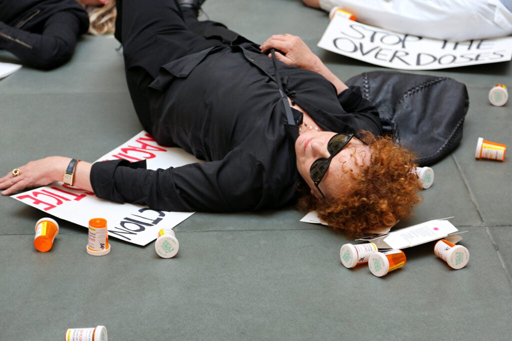 Nan Goldin makaa maassa, ympärillä oransseja pilleripurkkeja ja mielenosoituskylttejä.
