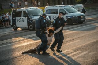 Antti Yrjösen valokuvassa kaksi poliisia raahaa mielenosoittajaa tien yli. Taustalla poliisiautoja ja ihmisiä.