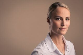 Kiira Korpi katsoo kameraan hymyillen, taustalla tasainen, beigenvärinen seinä.