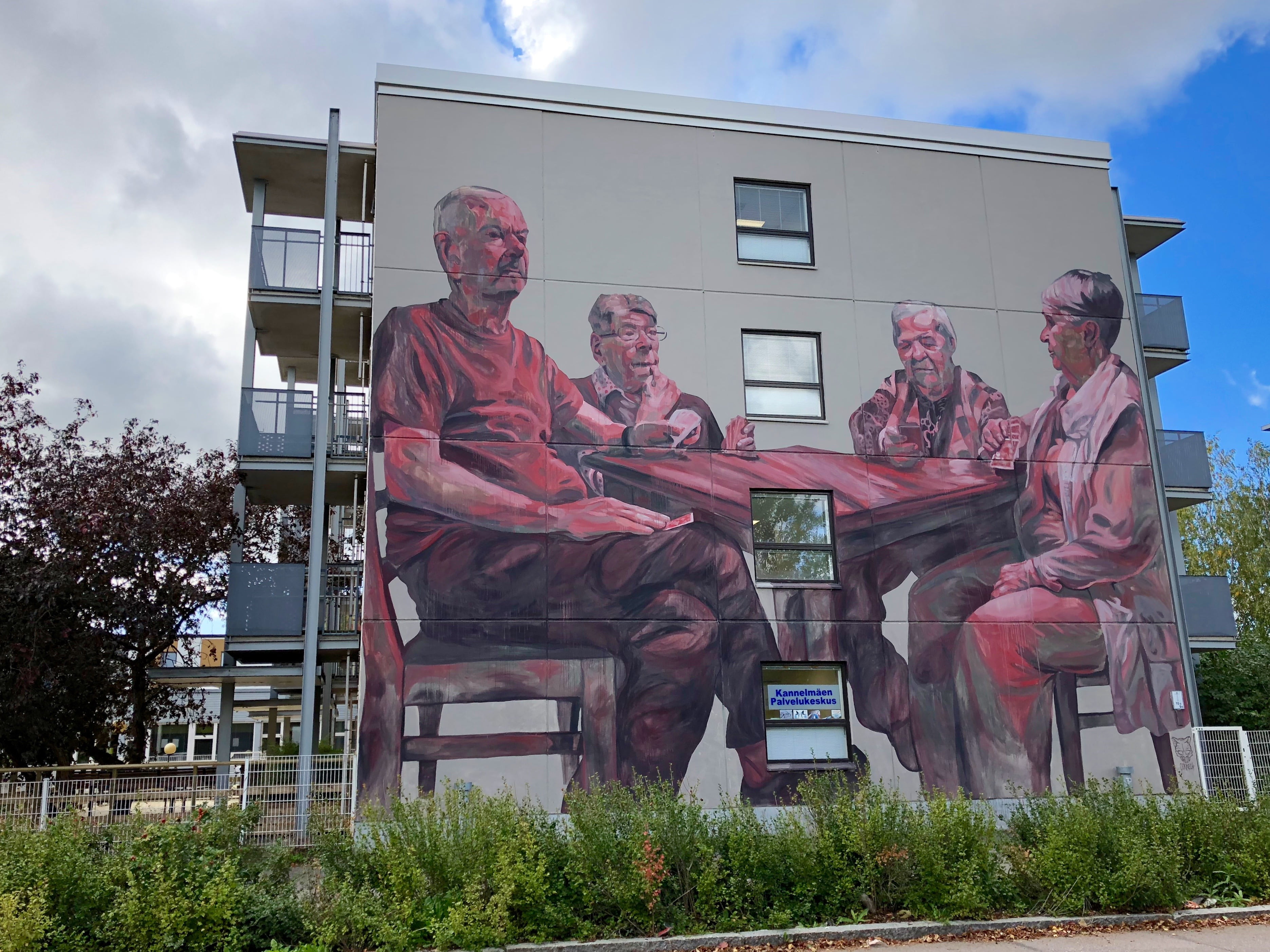 Milu Correchin muraali Kannelmäen palvelutalon seinässä. Muraalissa neljä iäkästä henkilöä istuu pöydän ääressä.