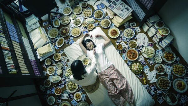 Nainen syöttää toista lattialla makaa naista. Ympärillä lukuisia lautasia ruokaa.