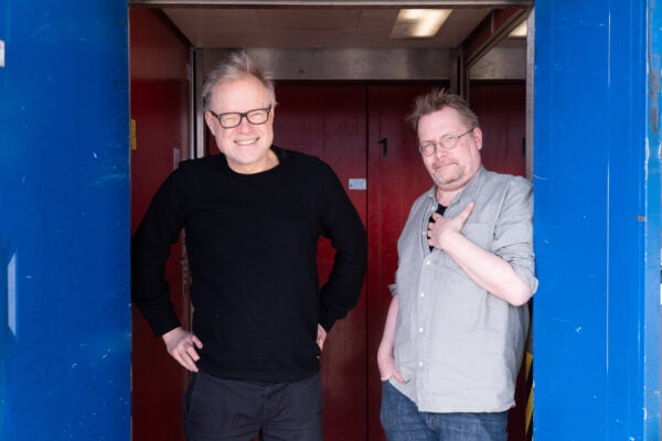 Esa Kirkkopelto ja Tuomas Rantanen hississä sinisten ovien välissä punaista taustaa vasten.