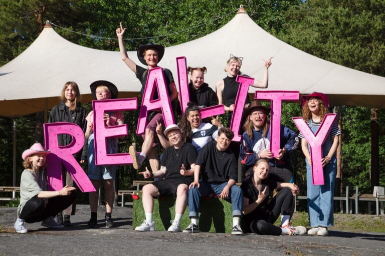 Joukko nauravia ihmisiä, pitelevät käsissään kirjaimia, jotka muodostavat sanan "REALITY".