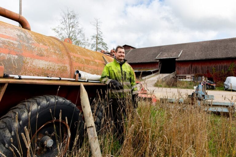 Kyrö nojaa lannoituslaitteeseen, taustalla maatalousrakennus.