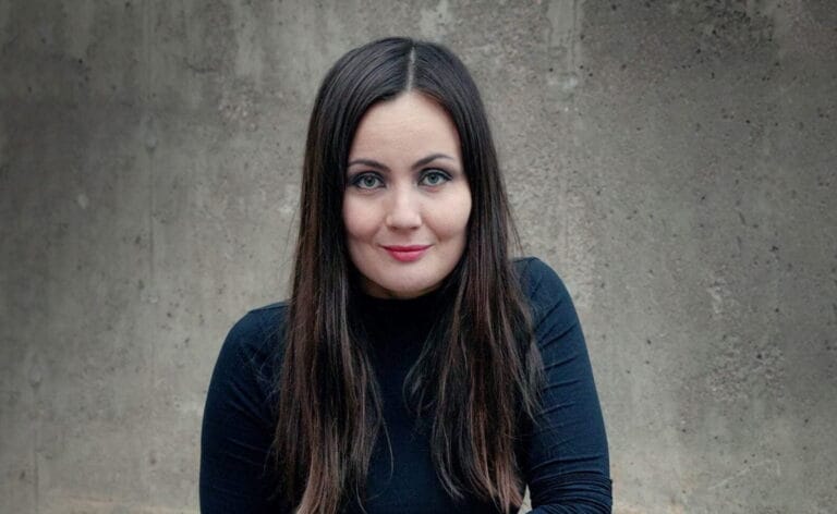 Emilia Kukkala katsoo kameraan hymyillen, taustalla harmaa seinä.