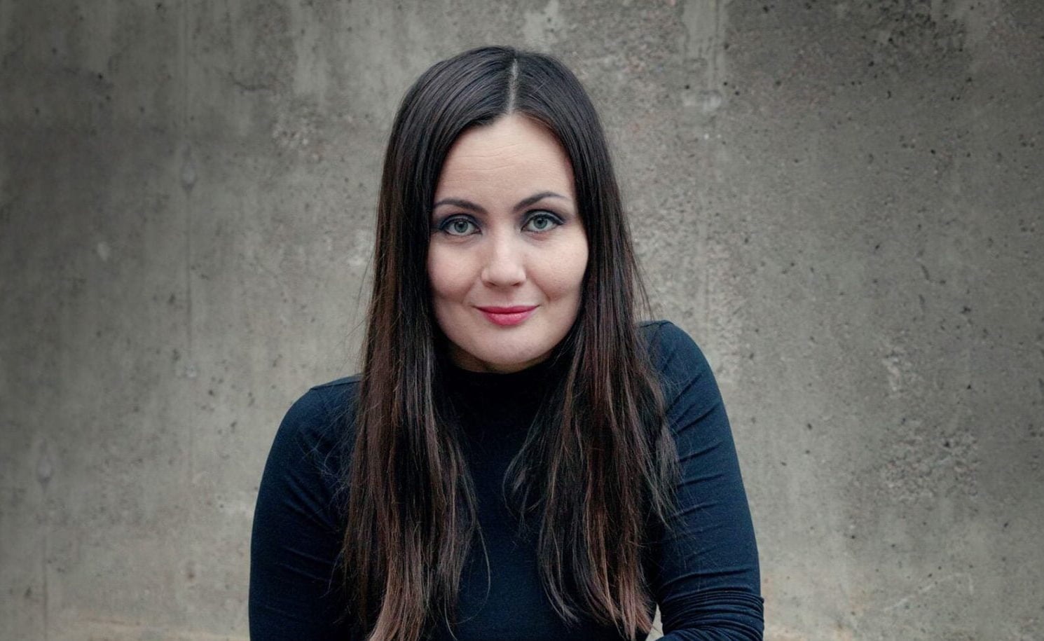 Emilia Kukkala katsoo kameraan hymyillen, taustalla harmaa seinä.