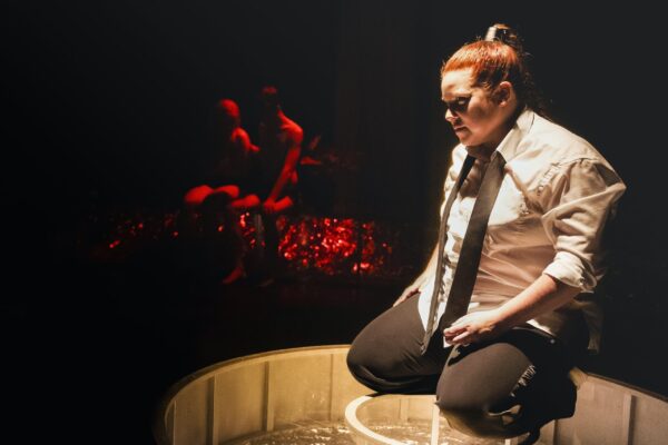 Teatteriesitys syntiinlankeemuksesta tuo esiin Saatanan oman tragedian Saatanaa esittävä Riikka Papunen polvistuneena kohdennusvalon alla. Taustalla punasävyiset Aatamin ja Eevan hahmot.