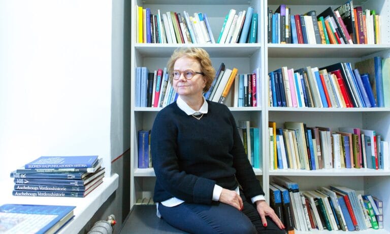 Kolbe istuu penkillä, katsoo pois kamerasta, taustalla kirjahylly.