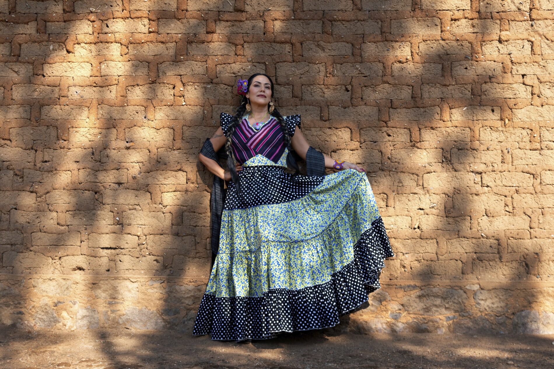 Lila Downs katsoo kameraan hymyillen, pitelee hameensa helmaa, taustalla tiiliseinä.