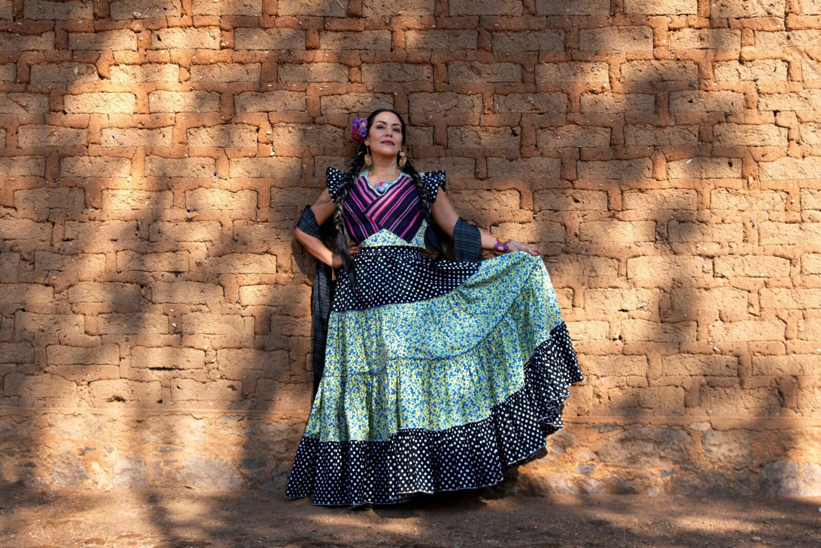 Lila Downs katsoo kameraan hymyillen, pitelee hameensa helmaa, taustalla tiiliseinä.