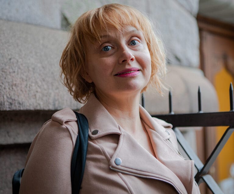 Maria Säkö katsoo kameraan. Päällään vaalea takki, takana kiviseinä.