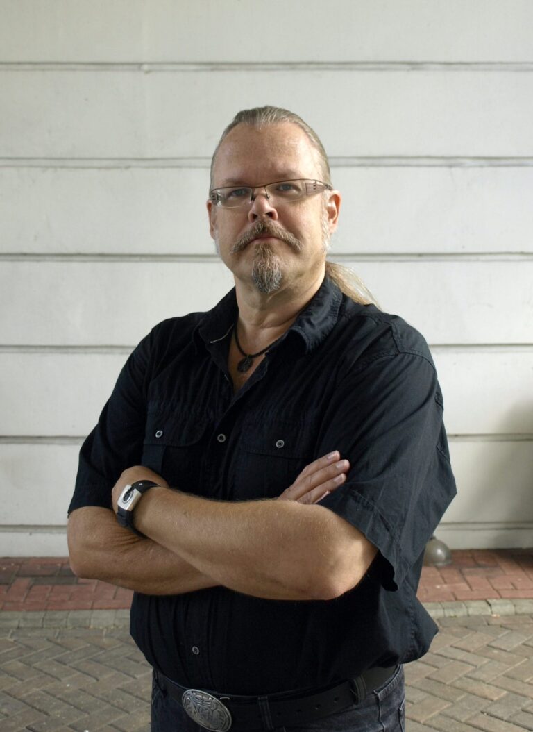 Markus Hotakainen katsoo kameraan, kädet puuskassa, tausatlla harmaa seinä.