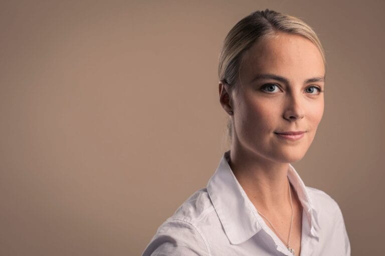 Kiira Korpi katsoo kameraan hymyillen, taustalla tasainen, beigenvärinen seinä.
