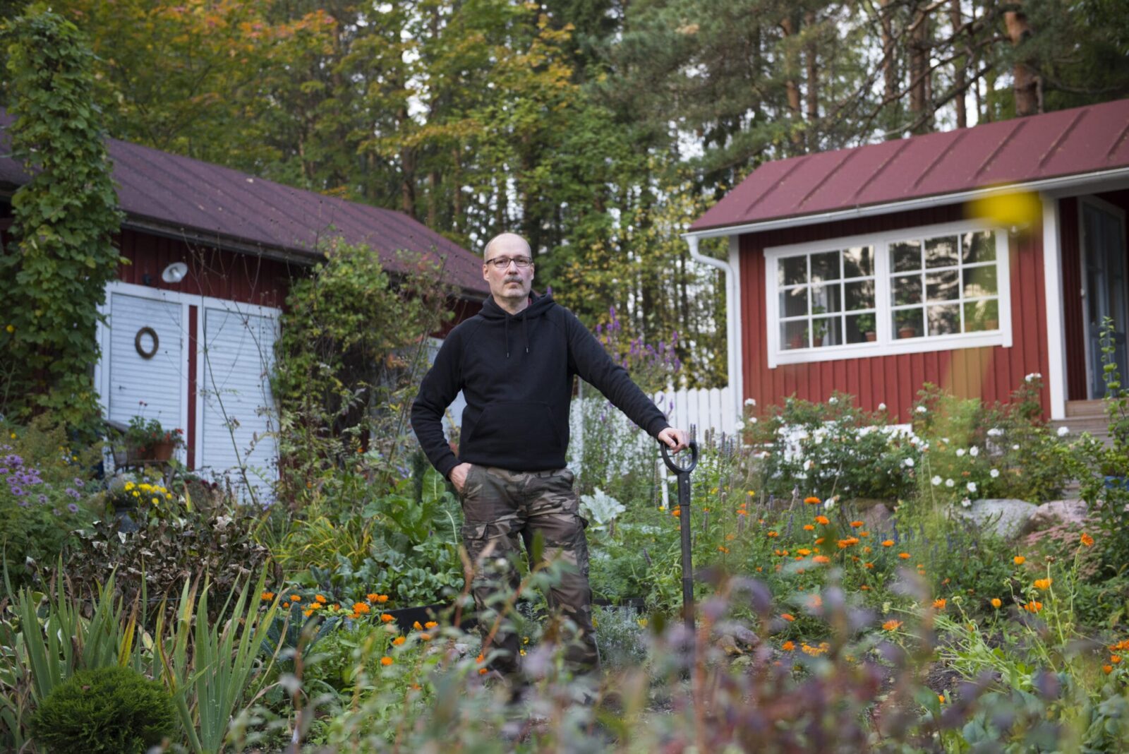 Kärkkäinen puutarhassa nojaamassa lapioon. Taustalla kaksi ulkorakennusta.