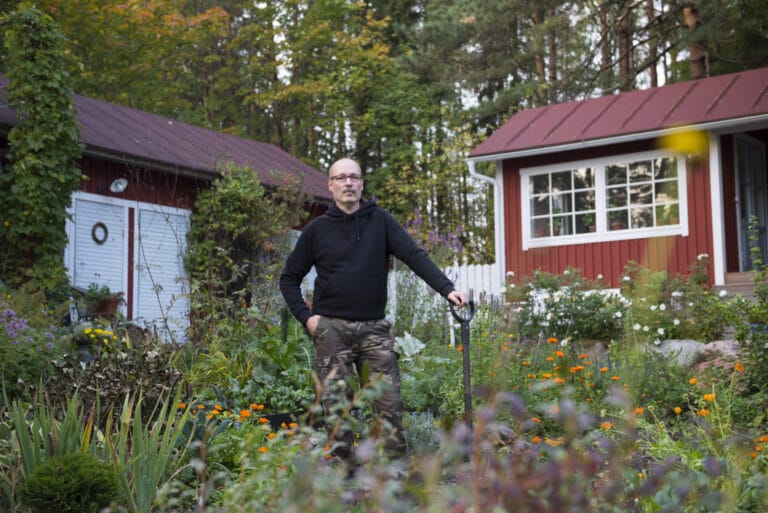 Kärkkäinen puutarhassa nojaamassa lapioon. Taustalla kaksi ulkorakennusta.