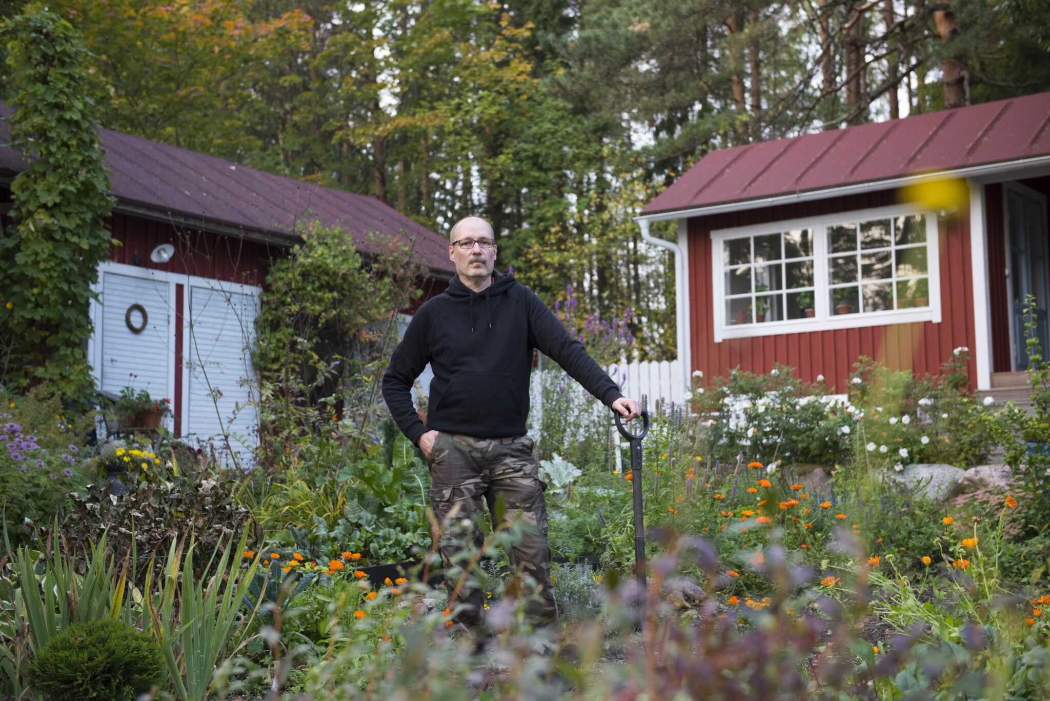 Kärkkäinen puutarhassa nojaamassa lapioon. Taustalla kaksi ulkorakennusta.