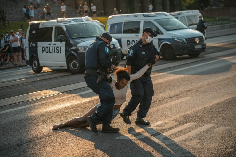 Antti Yrjösen valokuvassa kaksi poliisia raahaa mielenosoittajaa tien yli. Taustalla poliisiautoja ja ihmisiä.