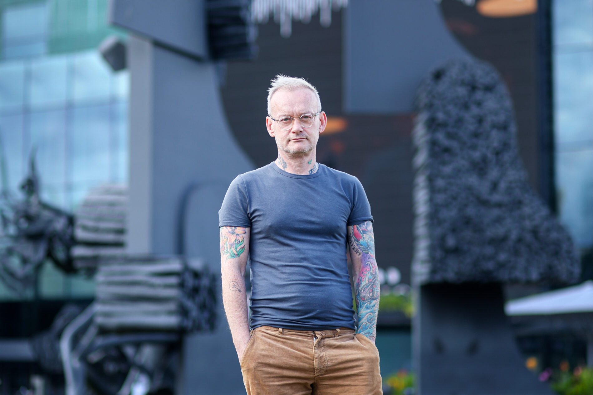 Conor Mitchell katsoo vakavalla ilmeellä kameraan tumman metallisen patsaan edessä rennosti seisoen, tatuoidut kädet housujen taskuissa.