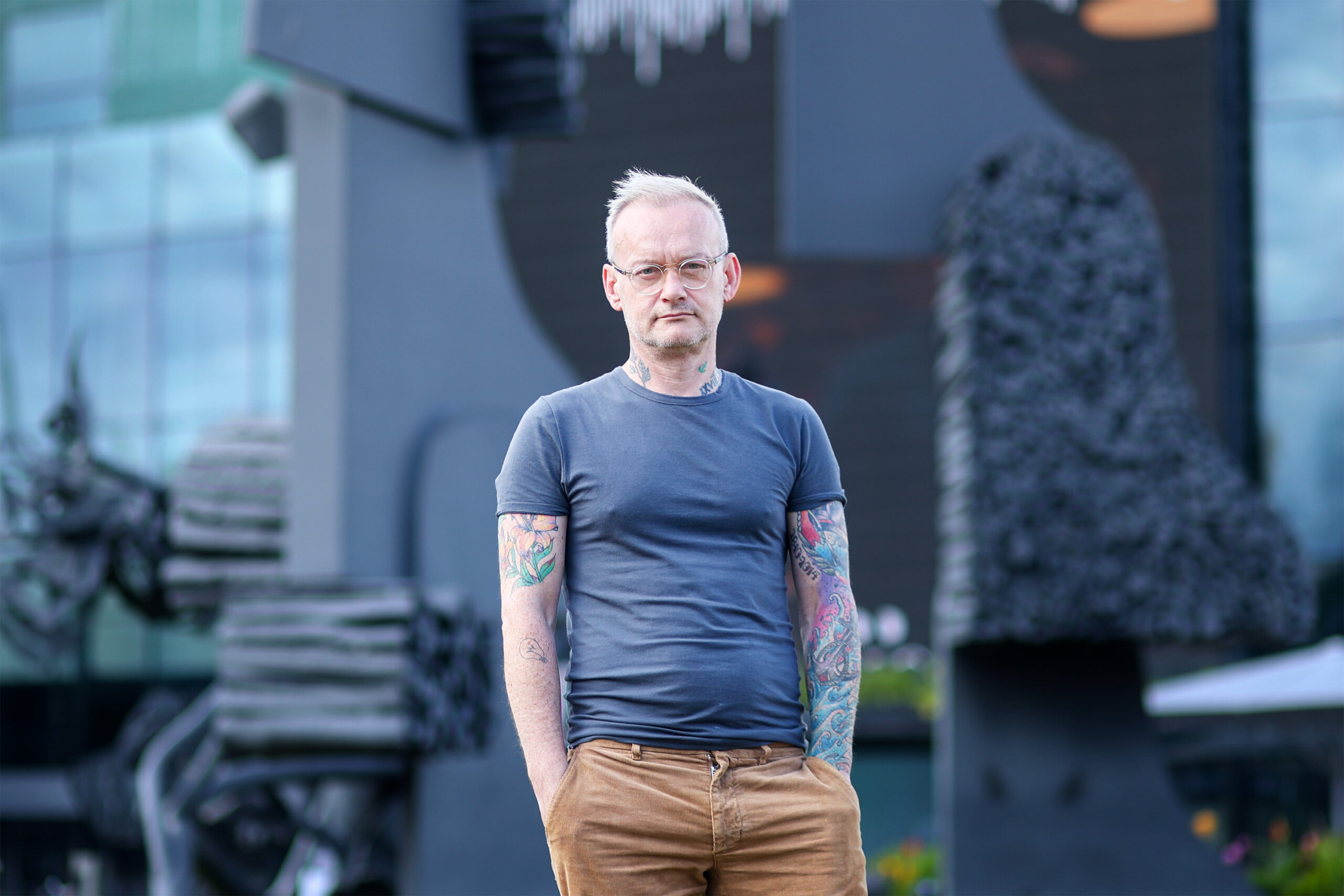 Conor Mitchell katsoo vakavalla ilmeellä kameraan tumman metallisen patsaan edessä rennosti seisoen, tatuoidut kädet housujen taskuissa.